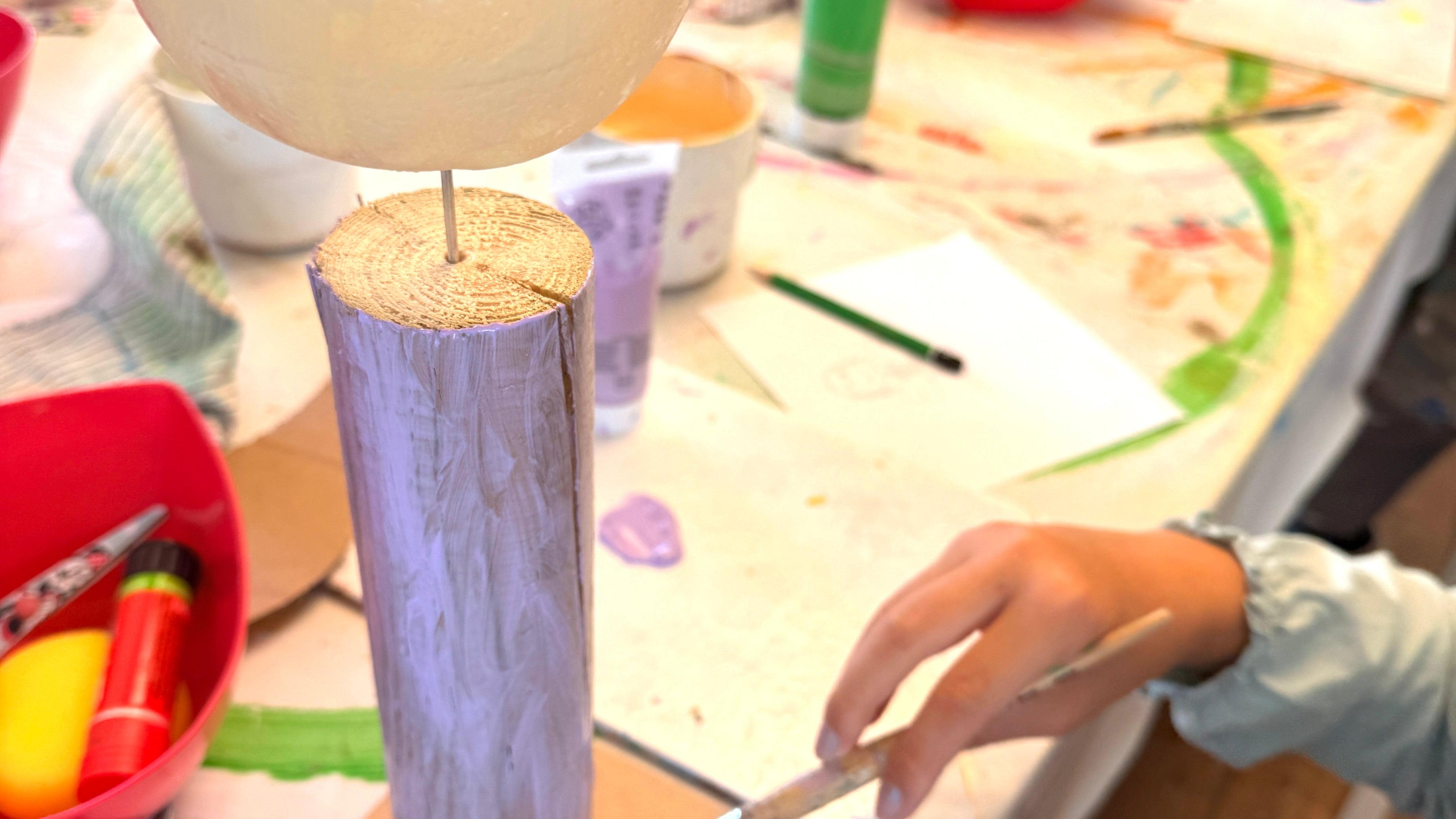 Eine Hand eines Kindes bemalt einen Holzfiguren-Rohling mit einem Pinsel und lila Farbe an einem kreativen Basteltisch der PINSELTANTE Akademie.