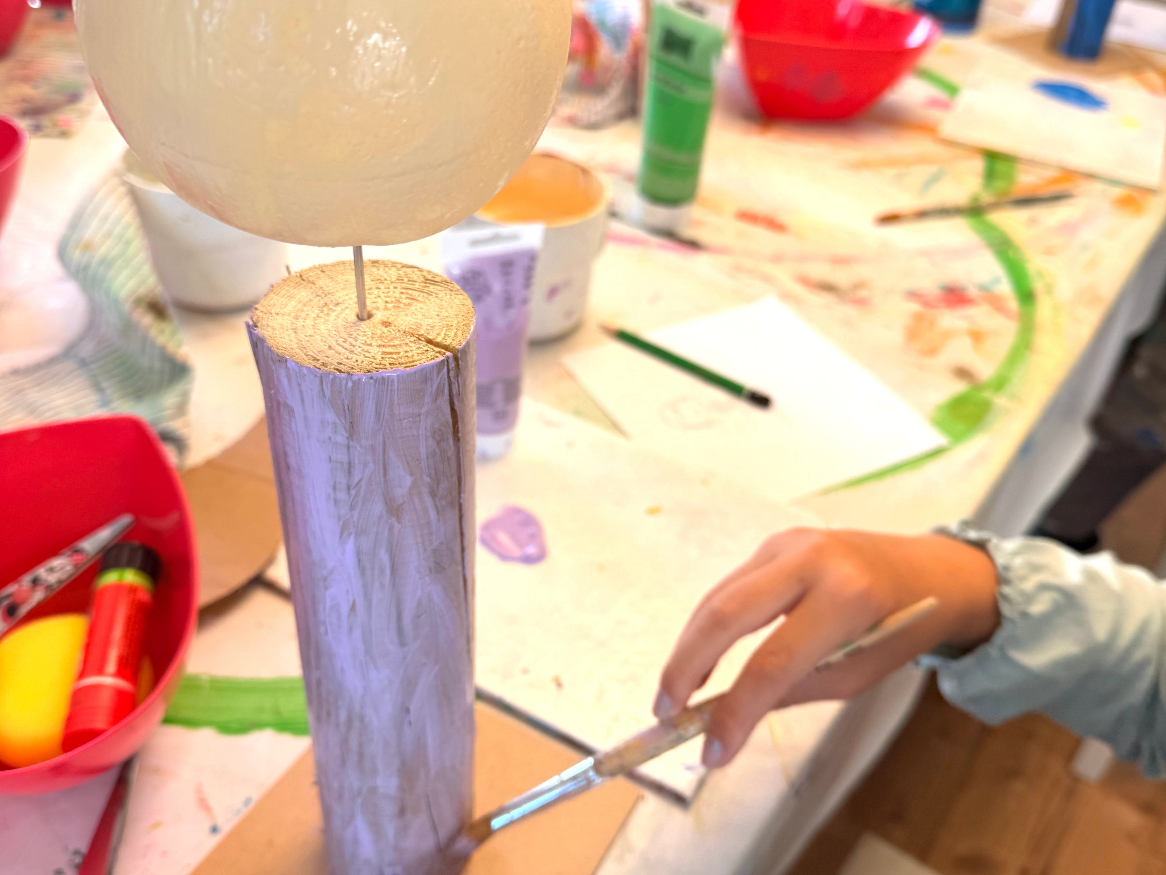Eine Hand eines Kindes bemalt einen Holzfiguren-Rohling mit einem Pinsel und lila Farbe an einem kreativen Basteltisch der PINSELTANTE Akademie.
