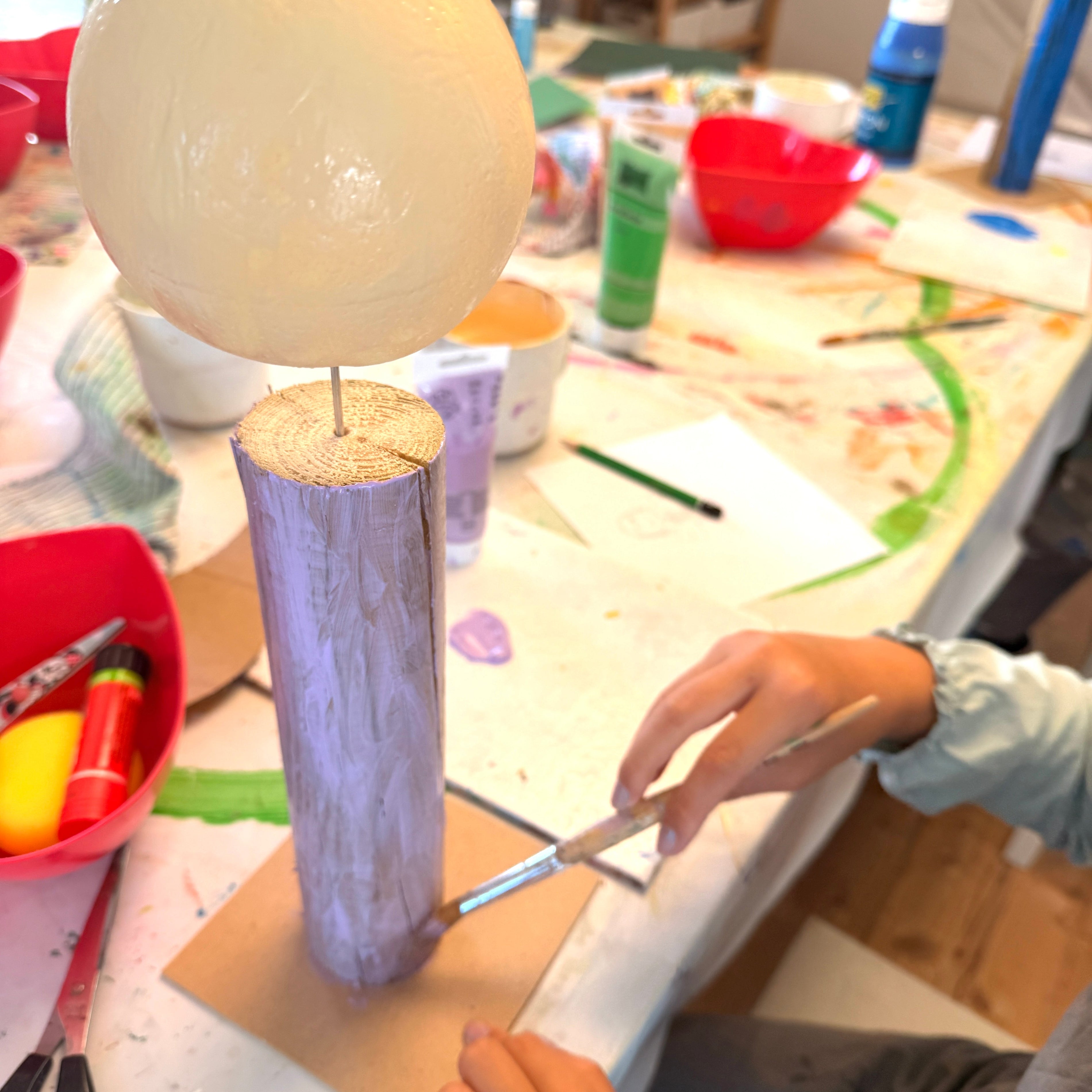 Eine Hand eines Kindes bemalt einen Holzfiguren-Rohling mit einem Pinsel und lila Farbe an einem kreativen Basteltisch der PINSELTANTE Akademie.